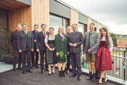 Eine Gruppe von Menschen in traditioneller Kleidung steht lächelnd für ein Foto auf einem Balkon. Die Umgebung ist ein modernes Gebäude mit Glasfenstern und einer Holzwand.