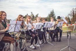 Eine Gruppe von Kindern und jungen Erwachsenen sitzt in einer Reihe und spielt Blasinstrumente, darunter Flöten und Klarinetten. Sie tragen formelle Kleidung und halten Notenblätter. Im Hintergrund ist ein unscharfer Park mit Bäumen und einer Bank zu sehen.