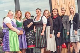 Eine Gruppe von Frauen in traditioneller deutscher Kleidung posiert für ein Foto vor einem Holzgebäude. Sie lächeln und stehen eng beieinander.