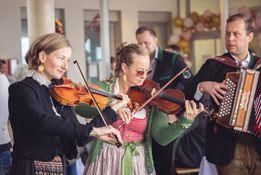 Drei Musiker spielen auf der Bühne. Zwei Frauen spielen Geige und ein Mann spielt Akkordeon. Sie befinden sich in einer festlichen Umgebung, wahrscheinlich bei einer Feier.