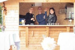 Drei Personen stehen hinter einer Bar in einem rustikalen Holzraum mit weißen Tischdecken. Ein Mann winkt mit beiden Händen, und zwei Frauen schauen zu. Auf dem Tresen befinden sich Flaschen und eine weiße Blume.