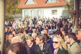 Eine große Menschenmenge versammelt sich vor einem Haus mit rotem Dach und weißen Wänden, viele in formeller Kleidung, möglicherweise für eine Hochzeit oder Feier.
