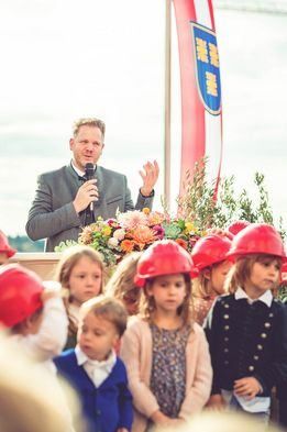 Ein Mann spricht in ein Mikrofon an einem Podium mit Blumen. Kinder in Schutzhelmen lauschen. Eine Fahne mit einem Logo ist im Hintergrund.