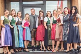 Eine Gruppe von Frauen und einem Mann in traditioneller deutscher Kleidung steht vor einem Gebäude und posiert für ein Foto. Die Frauen tragen farbenfrohe Kleider mit Schürzen, und der Mann ist in einem Anzug.