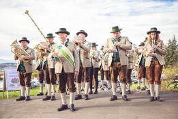 Eine Gruppe von Musikern in traditioneller bayerischer Kleidung, einschließlich grüner Hüte und brauner Hosen, spielt verschiedene Instrumente wie Trompeten, Posaunen und Tuben an einem sonnigen Tag.