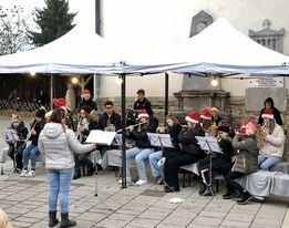 Eine Gruppe von Musikern mit Weihnachtsmützen spielt unter einem weißen Zelt, mit einem Dirigenten vor ihnen und Instrumenten auf dem Tisch.