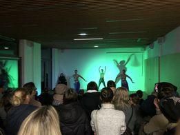 Eine Gruppe von Menschen beobachtet eine Aufführung auf der Bühne mit grünem Licht, an der drei Darsteller teilnehmen. Das Publikum sitzt im Halbkreis und hält die Momente mit ihren Kameras fest.