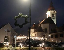 Eine Kirche mit Turm und Spitze ist nachts beleuchtet. Ein Kranz hängt an einem Pfosten vor der Kirche. Mehrere Autos sind vor der Kirche geparkt.