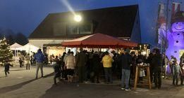 Ein nächtlicher Markt ist voller Menschen, einige in Jacken und Mützen, unter einem roten Baldachin. Ein Weihnachtsbaum und eine Schneemann-Statue sind im Hintergrund zu sehen.