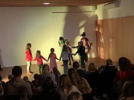 Mehrere Kinder halten sich an den Händen und tanzen auf einer Bühne vor einem Publikum. Die Bühne ist von einem Licht beleuchtet und die Wände sind weiß gestrichen.