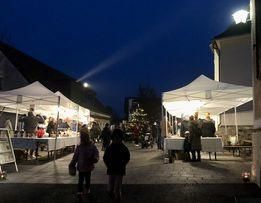 Eine nächtliche Marktszene mit mehreren weißen Zelten und leuchtenden Laternen. Menschen sind um Tische versammelt, einige stehen und andere sitzen. Die Straße ist schwach beleuchtet von einer Straßenlaterne.