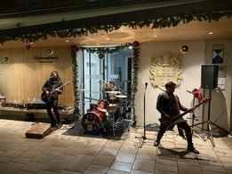 Eine Band spielt auf einer gefliesten Bühne vor einem Gebäude mit Weihnachtsdekoration. Eine Person spielt Gitarre, eine andere Schlagzeug und die dritte Person singt in ein Mikrofon.