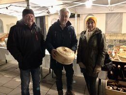Drei Personen stehen zusammen unter einem Zelt, zwei Männer und eine Frau. Der Mann in der Mitte hält einen großen runden Käse. Sie alle lächeln. Hinter ihnen steht ein Tisch mit verschiedenen Gegenständen darauf.