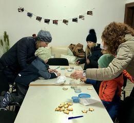 Mehrere Personen versammeln sich um einen Tisch mit Keksen und Bastelutensilien. Eine Frau in einer blauen Mütze beugt sich über ein Kind im Rollstuhl, und eine Frau in einer beigen Jacke reicht einem anderen Kind einen Keks. An der Wand hängen gerahmte Bilder und eine Topfpflanze steht in der Ecke.