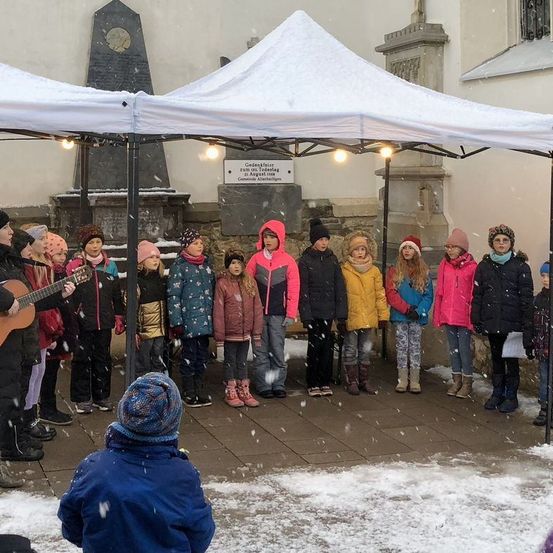 Eine Gruppe von Kindern steht unter einem Zeltdach im Schnee, in Winterkleidung und Mützen. Ein Kind spielt Gitarre.