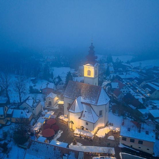 Luftaufnahme einer Kirche im Schnee, umgeben von winterlicher Landschaft mit Nebel. Gebäude sind beleuchtet, und ein Weihnachtsbaum steht davor.