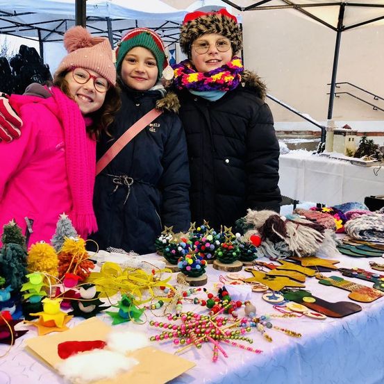 Drei Kinder lächeln vor einem Tisch voller selbstgemachter Weihnachtsdekorationen. Sie sind warm gekleidet, mit Schals und Mützen. Hinter ihnen steht ein weißes Zelt und eine Treppe.