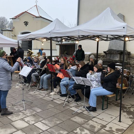 Ein Blechbläserensemble spielt unter einem Zelt, mit sitzenden und stehenden Musikern, und einem Dirigenten, der sie leitet. Eine Kirche ist im Hintergrund sichtbar.