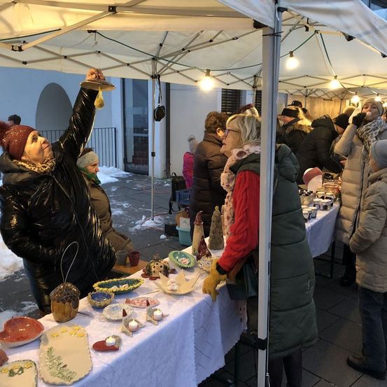 Ein Outdoor-Markt mit einem weißen Zelt. Menschen in Winterkleidung durchstöbern Gegenstände auf einem Tisch. Eine Frau hält ein kleines Objekt hoch, während eine andere Frau in Handschuhen den Tisch betrachtet.