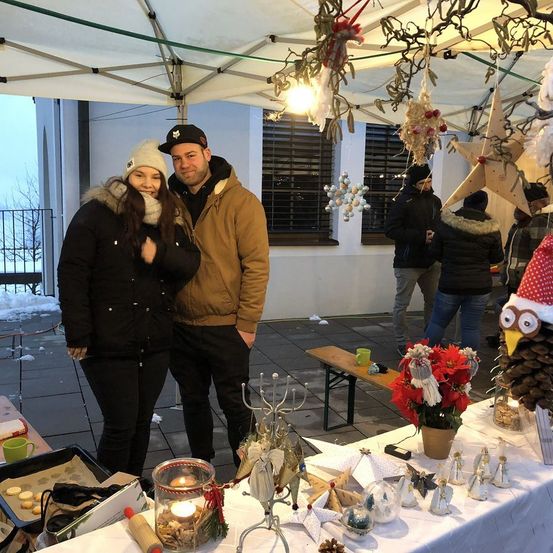 Ein Paar posiert für ein Foto auf einem Weihnachtsmarkt mit festlichen Dekorationen, Ornamenten und einem Tisch voller Gegenstände.