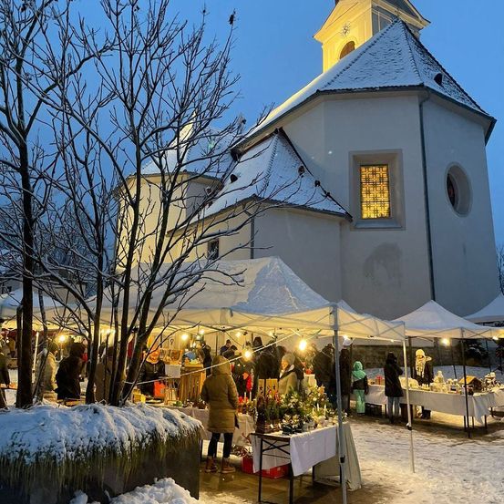 Ein Weihnachtsmarkt ist vor einer schneebedeckten Kirche aufgebaut. Zahlreiche Stände sind von Lichtern beleuchtet, und Menschen stöbern zwischen den ausgestellten Artikeln.