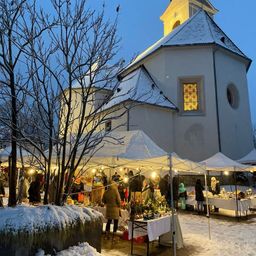 Ein Weihnachtsmarkt ist vor einer schneebedeckten Kirche aufgebaut. Zahlreiche Stände sind von Lichtern beleuchtet, und Menschen stöbern zwischen den ausgestellten Artikeln.