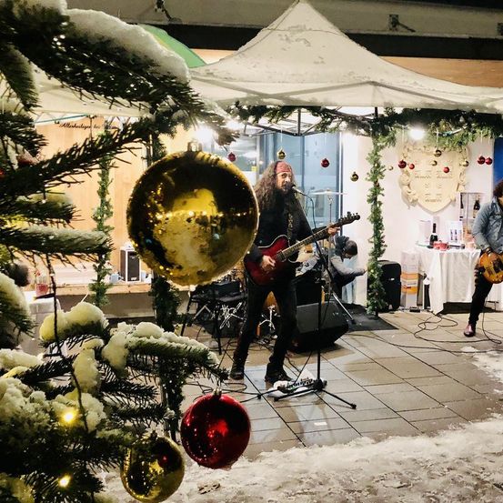 Zwei Musiker spielen auf einer verschneiten Bühne mit einem geschmückten Weihnachtsbaum. Der Lead-Gitarrist spielt eine rote Gitarre, während der andere E-Bass spielt. Im Hintergrund steht ein Zelt und ein Tisch mit Flaschen.