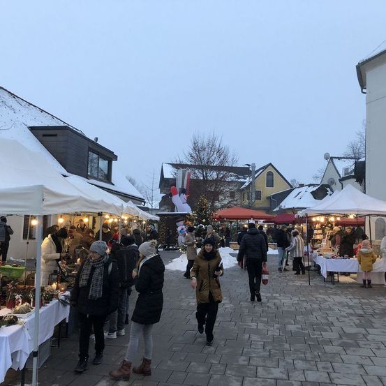 Ein Weihnachtsmarkt mit verschneiten Dächern und festlichen Dekorationen, Menschen gehen vorbei und kaufen an verschiedenen Ständen ein.