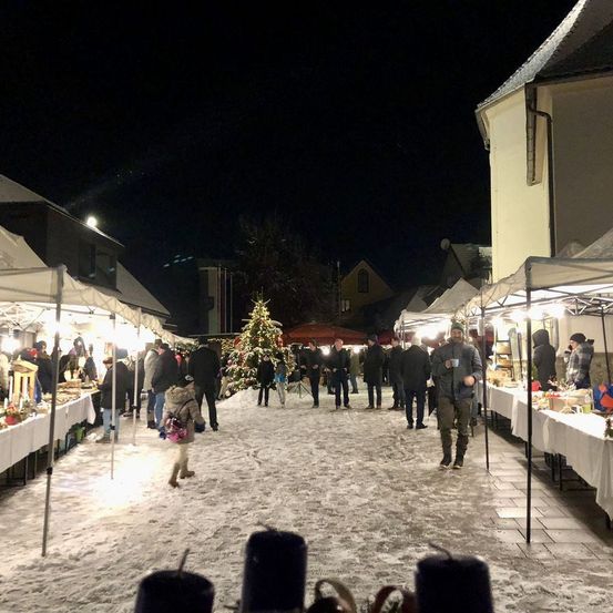 Ein Weihnachtsmarkt bei Nacht in einer verschneiten Stadt, mit einem Weihnachtsbaum in der Mitte und Leuten, die herumlaufen.
