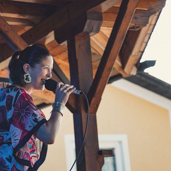 Eine Frau in einem farbenfrohen Oberteil und Ohrringen singt in ein Mikrofon. Sie steht unter einer Holzkonstruktion. Im Hintergrund befindet sich ein gelbes Haus mit weißen Fensterrahmen und einem Fenster.