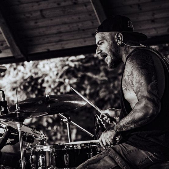 Ein Mann mit Tattoos auf den Armen spielt die Drums, hält Drumsticks, mit Mikrofonen und Becken vor ihm.