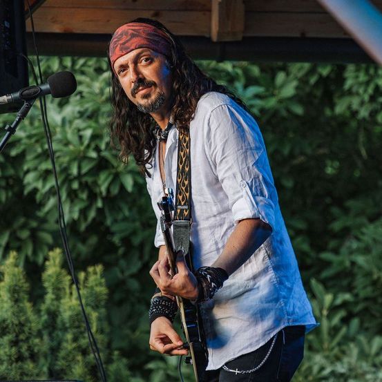 Ein Mann mit langen Haaren und einem Bandana spielt Gitarre auf der Bühne. Er trägt ein weißes Hemd und schwarze Hose. Vor ihm steht ein Mikrofon auf einem Ständer.