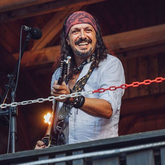 Ein Musiker steht auf der Bühne, lächelt und spielt Gitarre. Er trägt ein Stirnband, eine Halskette und eine Uhr. Vor ihm steht ein Mikrofon.