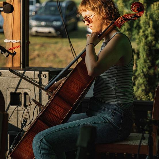 Eine Frau mit Sonnenbrille und einem ärmellosen Oberteil spielt Cello auf einem Stuhl im Freien. Vor ihr stehen ein Mikrofon und ein Lautsprecher. Im unscharfen Hintergrund ist ein geparktes Auto zu sehen.