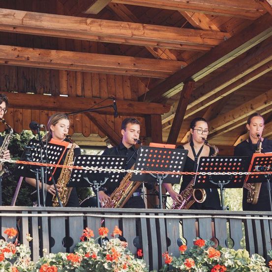 Eine Gruppe von Musikern in schwarzer Kleidung spielt auf einer Bühne mit einem Holzgeländer. Sie spielen Saxophon und stehen vor Notenständern. Die Bühne hat eine Holzdecke.