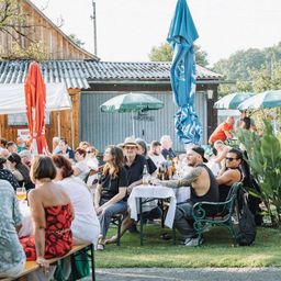 Eine Gruppe von Menschen versammelt sich in einem Außenbereich, wahrscheinlich für eine Party. Sie sitzen auf Stühlen um einen Tisch mit Flaschen. Einige tragen Hüte, Brillen und Sneakers. Der Bereich ist von Sonnenschirmen und Bäumen umgeben. Es gibt ein Haus mit einer Holzwand und einem Schild auf der linken Seite.