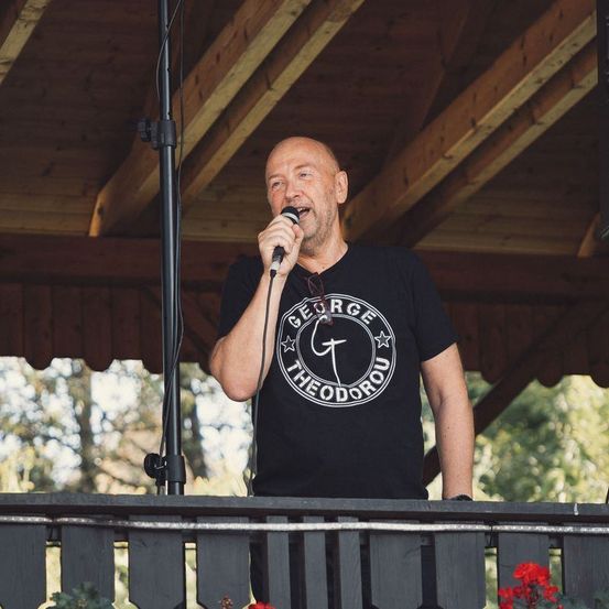 Ein kahlköpfiger Mann in einem George Theodorou T-Shirt steht auf einem Balkon, hält ein Mikrofon und singt. Der Balkon hat ein hölzerne Geländer und ist von Bäumen umgeben.