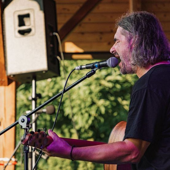 Ein Mann mit langen Haaren und Bart spielt Gitarre und singt in ein Mikrofon auf einer Bühne.