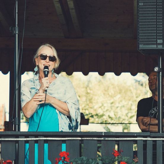 Eine ältere Frau mit Sonnenbrille und Ohrringen steht auf der Bühne mit einem Mikrofon und singt wahrscheinlich. Hinter ihr beobachtet ein Mann, und Lautsprecher sind aufgestellt.