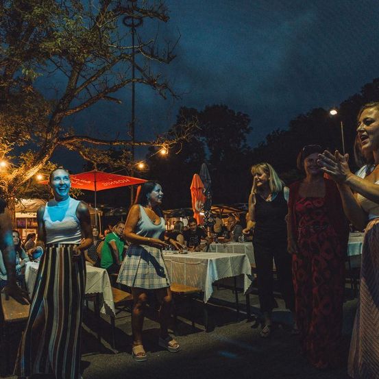 Freiluftessen mit mehreren tanzenden Frauen bei Nacht unter Lichterketten, Tischen und einem Regenschirm.