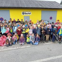 Eine Gruppe von Kindern und Erwachsenen posiert für ein Foto vor einem gelben Gebäude mit dem Schild Bauhof. Sie halten Stöcke, Taschen und einige tragen Hüte und Brillen.
