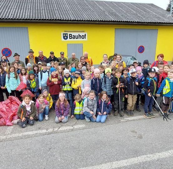 Eine Gruppe von Kindern und Erwachsenen posiert für ein Foto vor einem gelben Gebäude mit dem Schild Bauhof. Sie halten Stöcke, Taschen und einige tragen Hüte und Brillen.