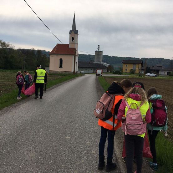 Schüler in Warnwesten gehen auf einer Landstraße in der Nähe einer Kirche und tragen Taschen. Dahinter ist ein Feuerwehrauto geparkt.