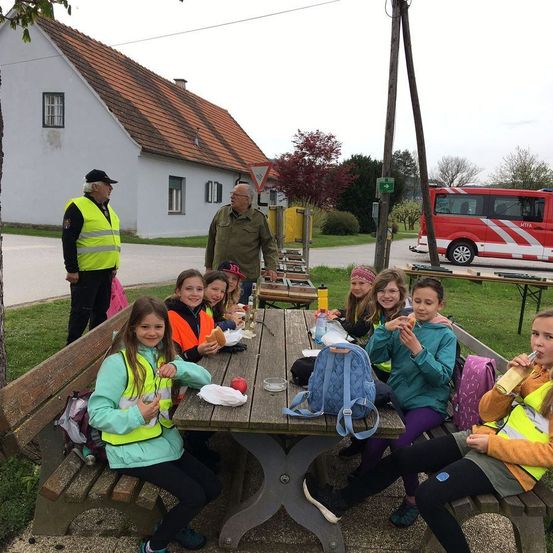 Eine Gruppe von Kindern in Sicherheitswesten genießt eine Mahlzeit an einem Picknicktisch im Freien. Zwei Erwachsene stehen in der Nähe, einer mit einer Kamera. Ein rotes Rettungsfahrzeug ist auf der Straße geparkt.
