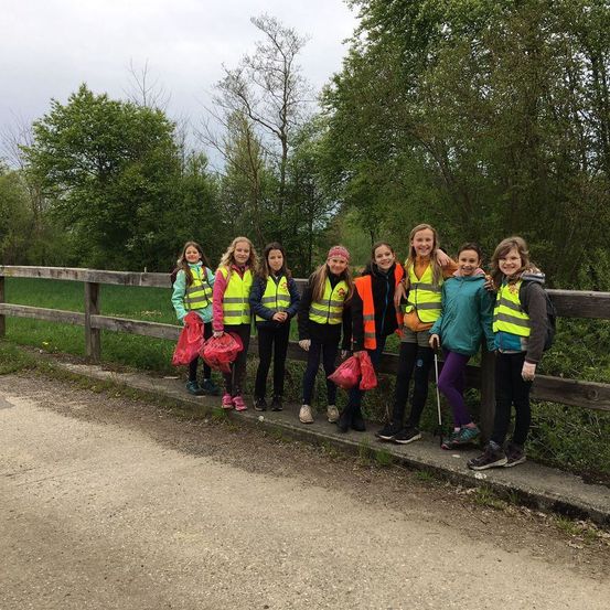 Eine Gruppe junger Personen in Sicherheitswesten posiert für ein Foto auf einer Brücke mit Taschen. Sie befinden sich in der Nähe eines Rasenbereichs mit Bäumen.