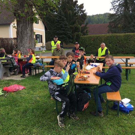 Eine Gruppe von Kindern und Erwachsenen sitzt auf Picknicktischen auf einer Rasenfläche und trägt reflektierende Westen. Sie essen mit Flaschen und Essen auf dem Tisch. Im Hintergrund befinden sich Häuser und Bäume.
