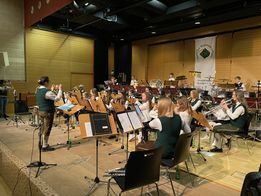 Ein großes Ensemble steht auf der Bühne, mit Musikern, die verschiedene Instrumente spielen, darunter Trompeten, Posaunen und Tuben. Ein Dirigent steht vorne und leitet die Aufführung. Die Musiker sind in grünen und weißen Uniformen gekleidet.