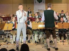 Ein Mann singt auf der Bühne, während ein anderer Mann neben ihm steht. Beide treten vor einem Orchester auf. Das Publikum beobachtet sie.