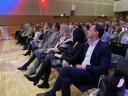 Ein überdachter Hörsaal mit Bühne hat ein Publikum aus formell gekleideten Männern und Frauen auf den Tribünen sitzend, die zur Bühne hinaufschauen.