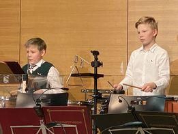 Zwei Jungen in einem Musikraum, einer mit einem Schlagzeug und der andere mit einer Marimba, treten auf. Sie sind von verschiedenen Musikinstrumenten und Ausrüstungsgegenständen umgeben.